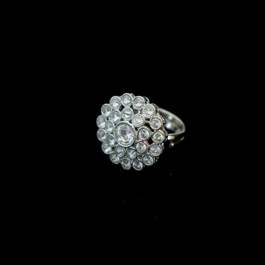 A silver-toned ring featuring a large, round floral cluster of clear gemstones, photographed on a dark reflective surface.