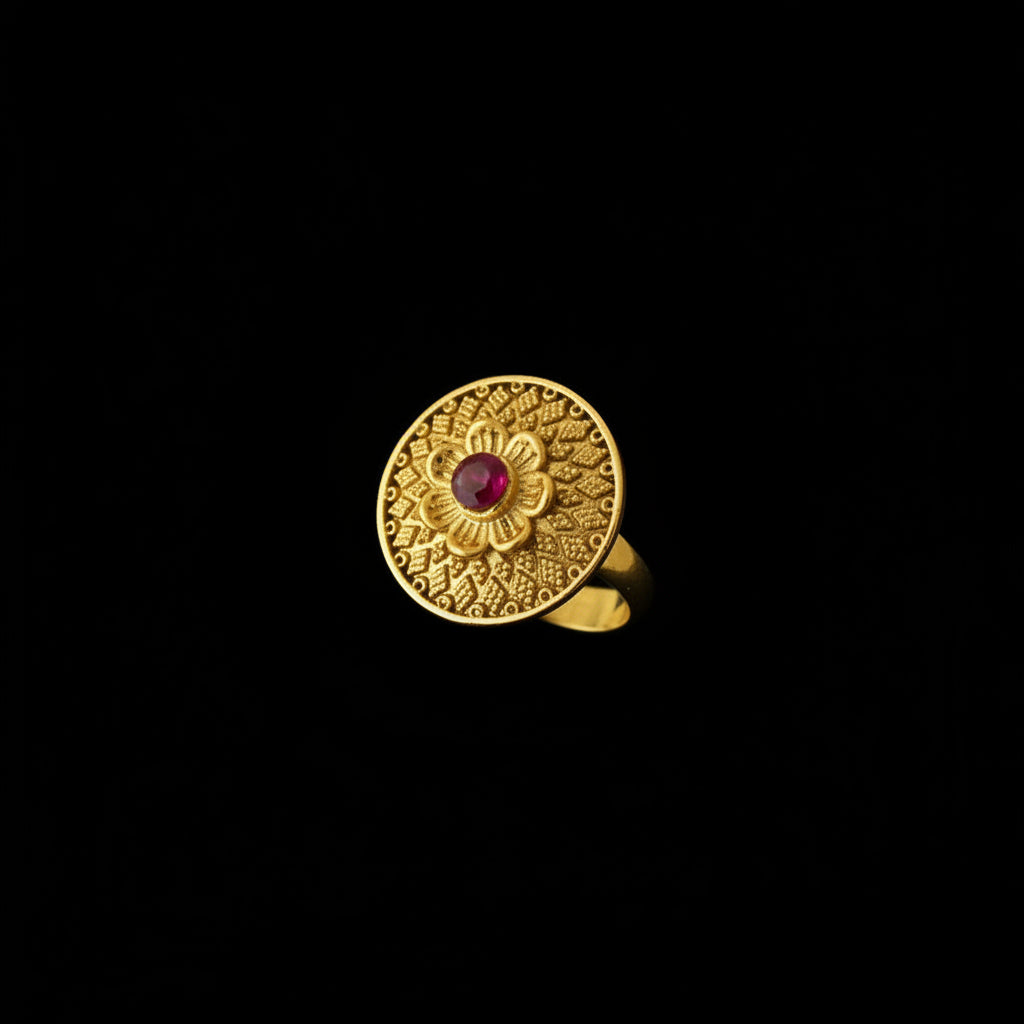A gold ring with an intricate circular filigree pattern and a small red center stone, displayed on a black hand.