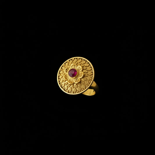 A gold ring with an intricate circular filigree pattern and a small red center stone, displayed on a black hand.
