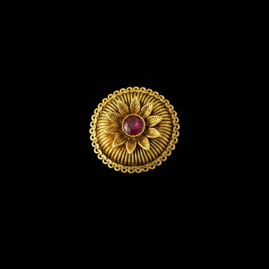 A gold ring with a circular sunburst design and a central red gemstone, displayed on a black gloved hand against a dark background.