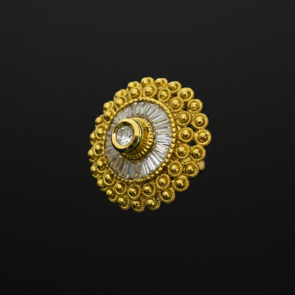A gold-toned traditional Indian ring with a circular filigree design, a central clear stone, and a white inlay, shown on a dark reflective surface. The SAHELI brand logo is visible in the top left corner.