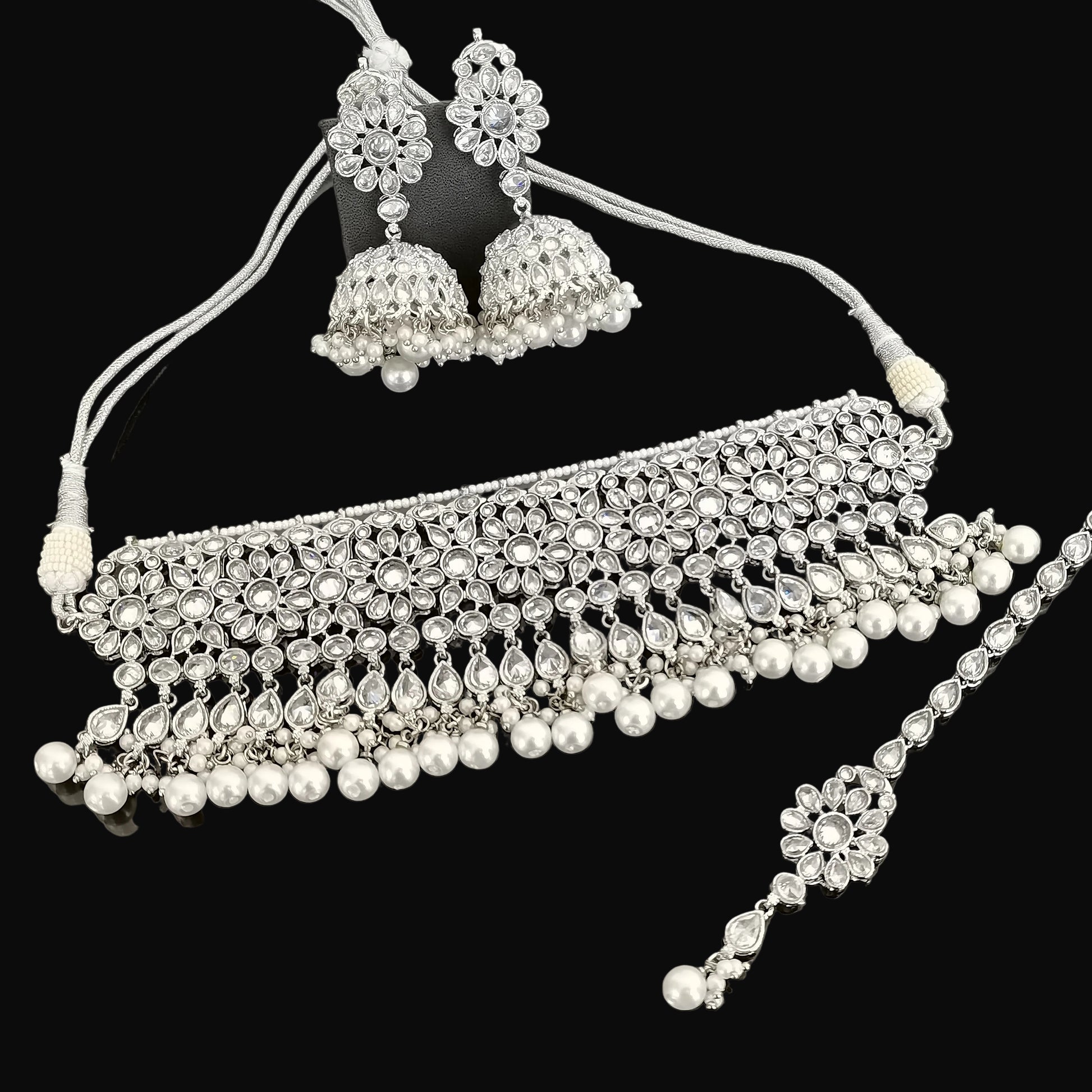 Silver jewelry set with necklace, earrings, and maang tikka on a dark surface with 'Saheli' branding.