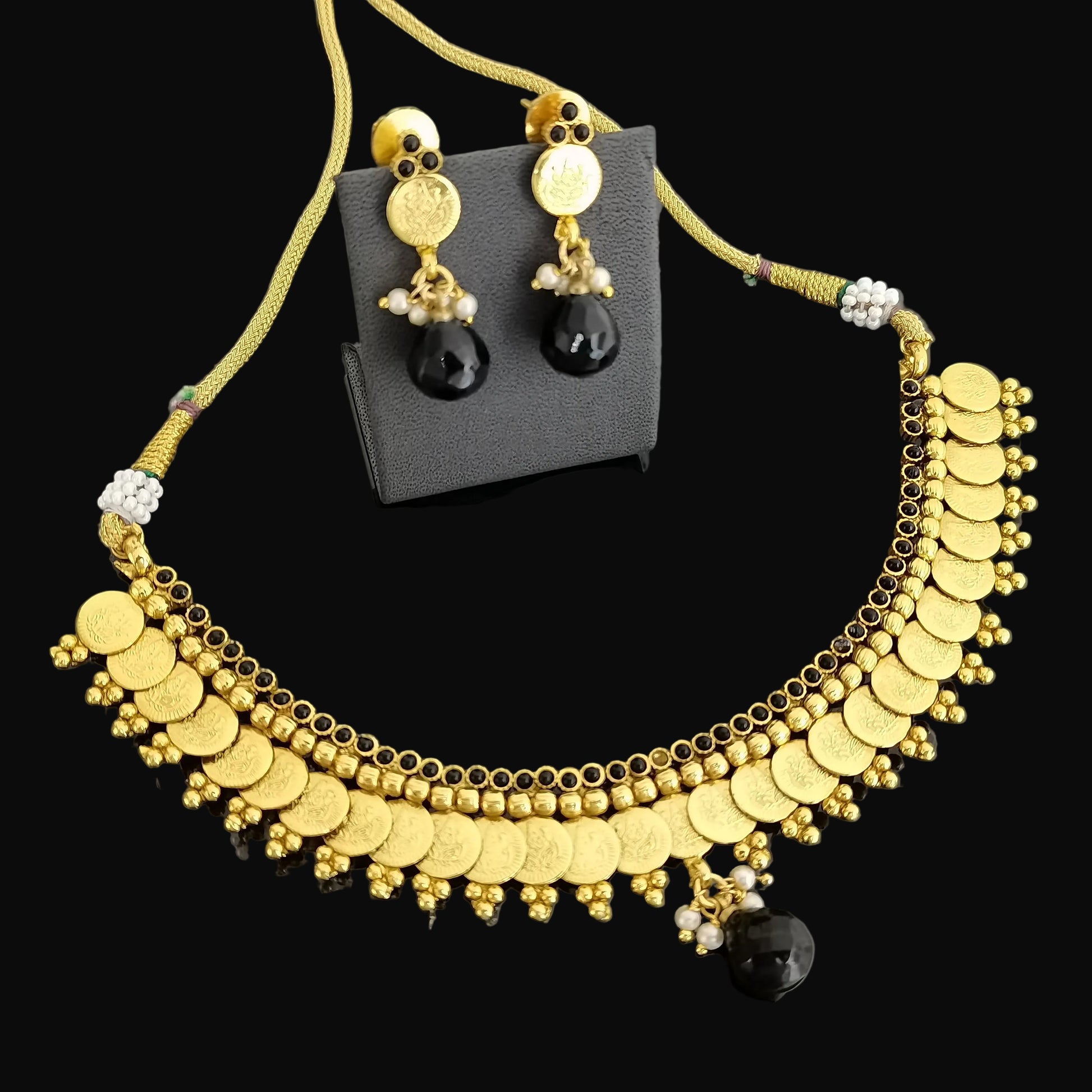 Gold coin choker necklace and matching drop earrings displayed on a black stand against a dark gray reflective background. The necklace features multiple rows of gold coins, beaded details, and a black bead pendant, with matching earrings shown on a black card.