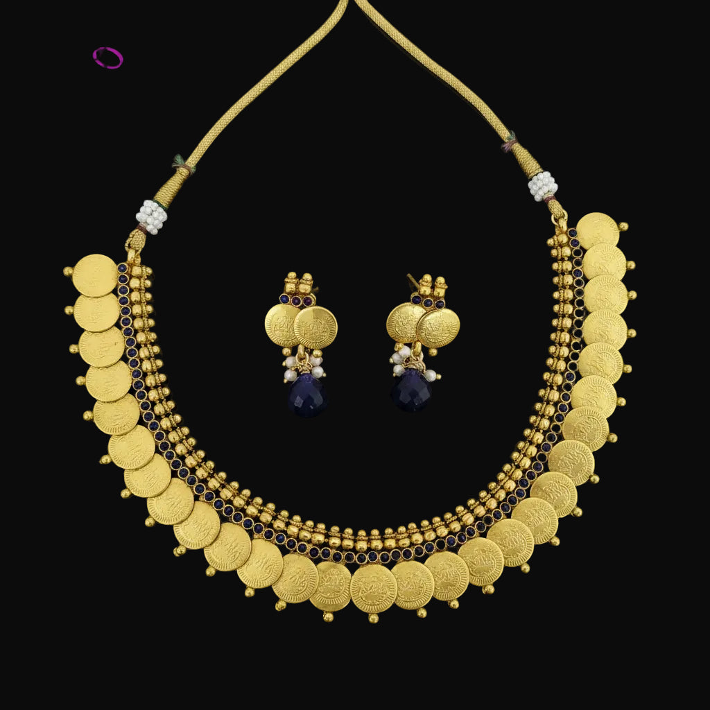 Gold coin necklace and matching drop earrings displayed on a dark reflective surface. The necklace features multiple rows of gold coins, small gold beads, and dark blue stone accents, with a gold cord closure.