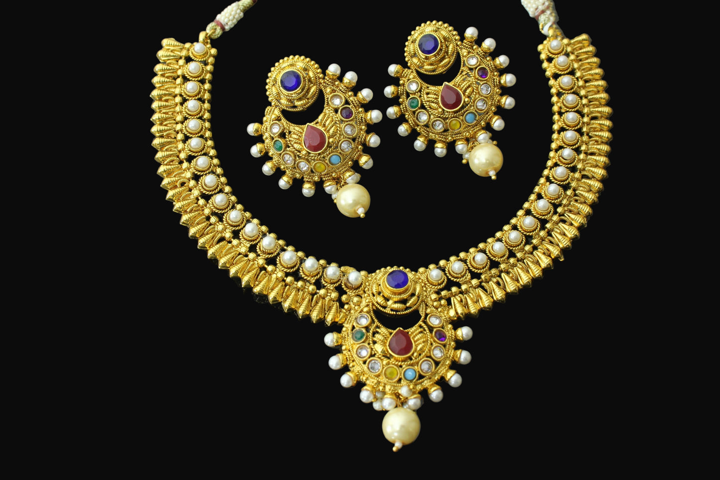 Gold necklace and earrings set with gemstones on a dark background, branded 'SAHELI'.