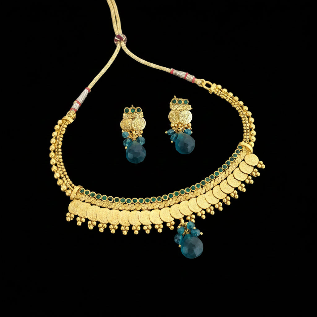 Gold-toned traditional necklace and matching drop earrings with green stone accents and blue teardrop beads, displayed on a dark reflective surface. Saheli My Best Friend logo appears in the top left.