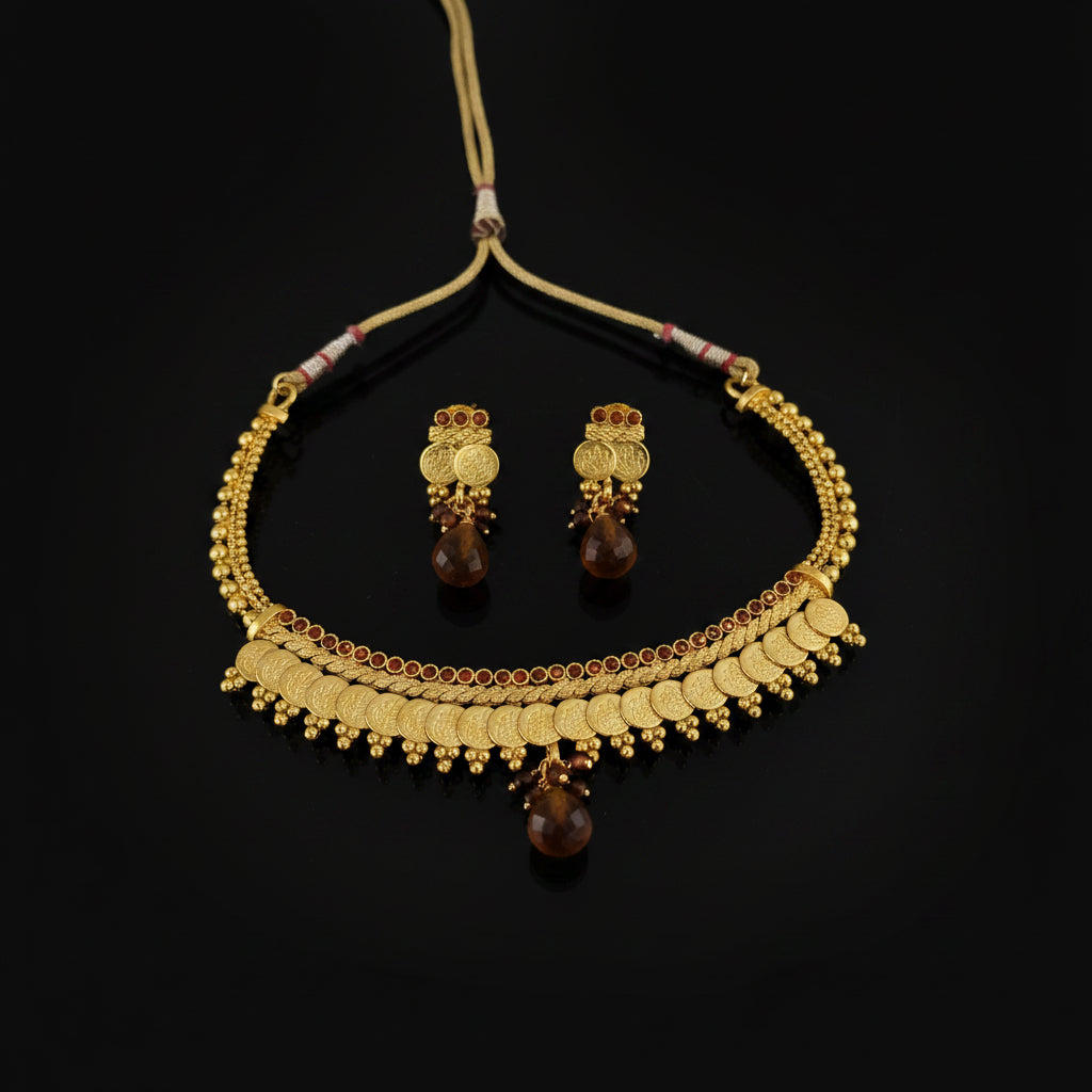 Gold-toned traditional necklace and matching earrings with amber-colored teardrop beads, displayed on a dark reflective surface. The necklace features gold coin-like disc elements, beaded borders, and small red gemstone accents.