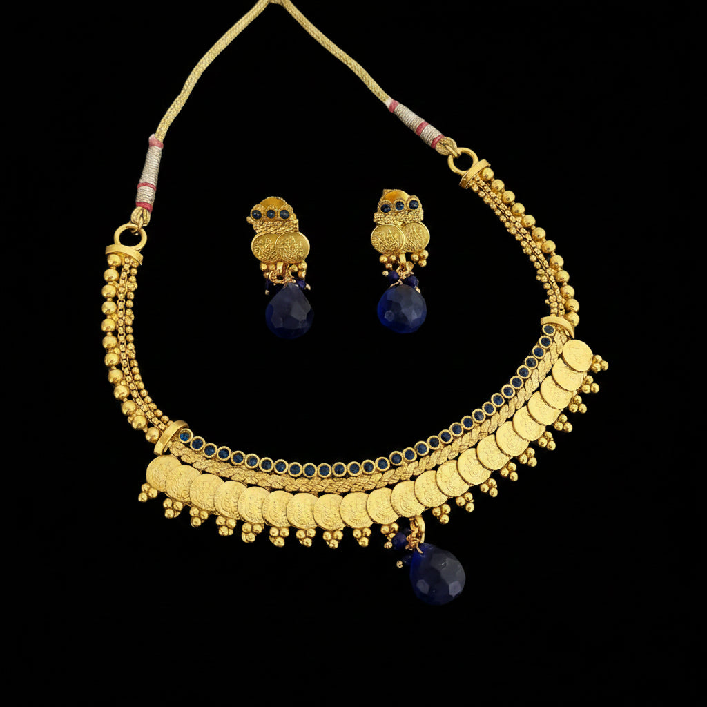 Gold-toned traditional necklace and matching owl-shaped earrings with dark blue teardrop stones, displayed on a dark gray reflective surface. The necklace has layered gold chains, coin-style discs, bead details, and dangling blue stones.