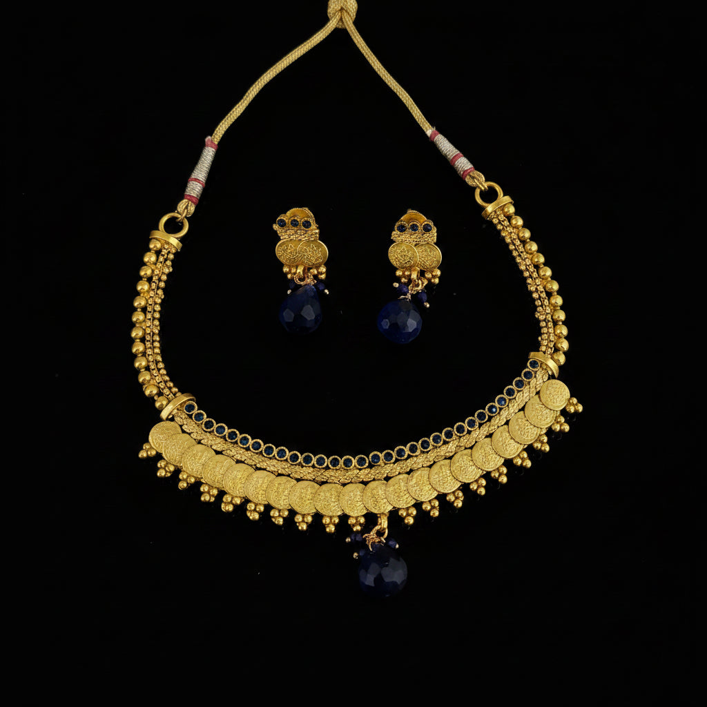 Gold-toned traditional necklace and matching owl-shaped earrings with dark blue teardrop beads, displayed on a dark gray reflective surface. The necklace has layered gold chains, coin-style discs, and dangling blue beads.