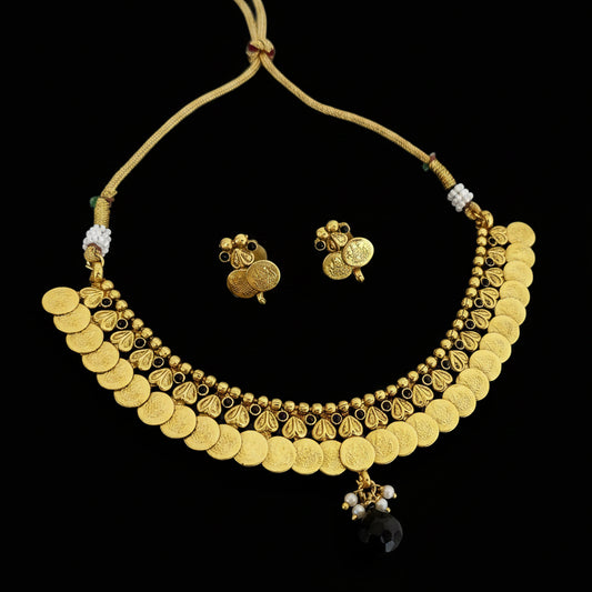 Gold coin necklace and matching stud earrings displayed on a dark reflective surface. The necklace features multiple rows of round gold coins, small bead details, and a dangling black faceted stone.
