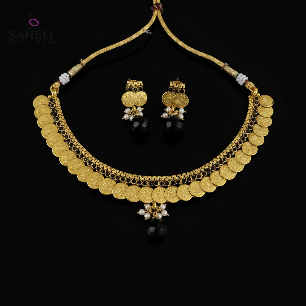 Gold coin necklace and matching earrings with black beads and white pearl accents, displayed on a dark reflective surface. The SAHeli by Saheli brand logo is visible in the top left corner.