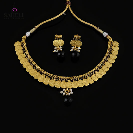 Gold coin necklace and matching earrings with black beads and white pearl accents, displayed on a dark reflective surface. The SAHeli by Saheli brand logo is visible in the top left corner.