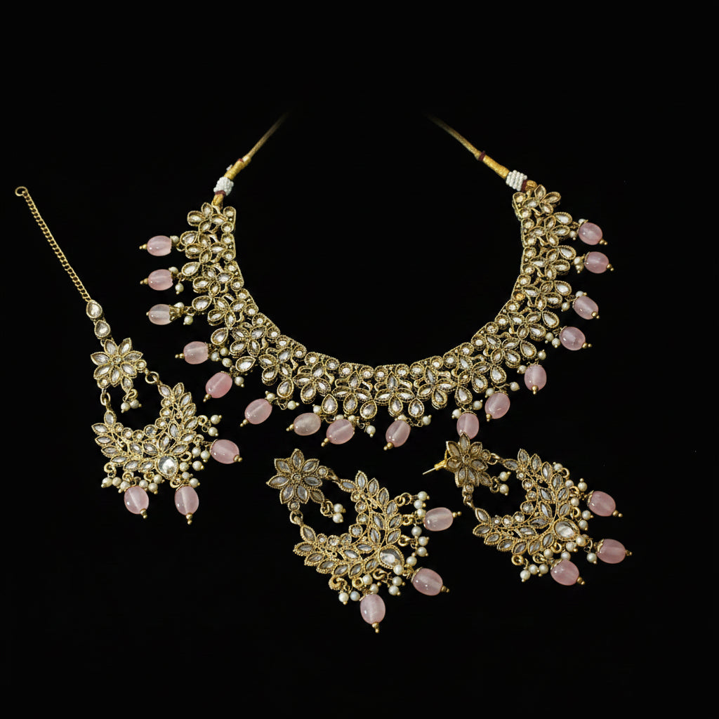 Gold-plated Indian ethnic jewelry set featuring a statement necklace, matching earrings, and a maang tikka, embellished with pink stones and white faux pearls, displayed on a dark reflective surface with the brand name 'SAHELL' visible in the background.