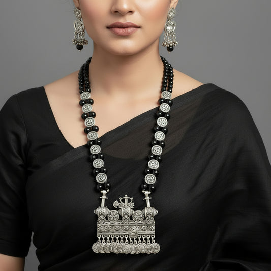 Black beaded necklace with silver pendant and earrings on a gray background, featuring the brand 'Saheli'.