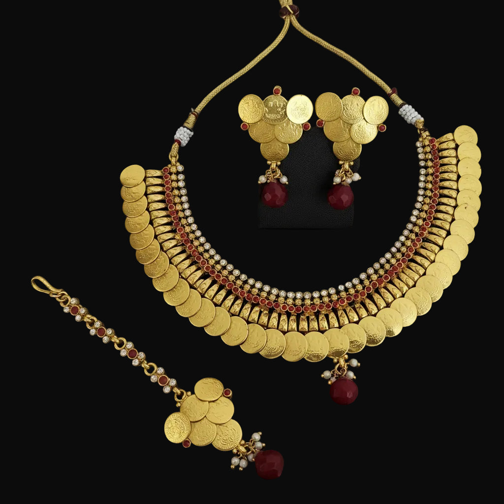 Gold coin necklace set with matching earrings and a maang tikka, featuring gold coins, red beads, and white pearl-like accents, displayed on a dark reflective surface.