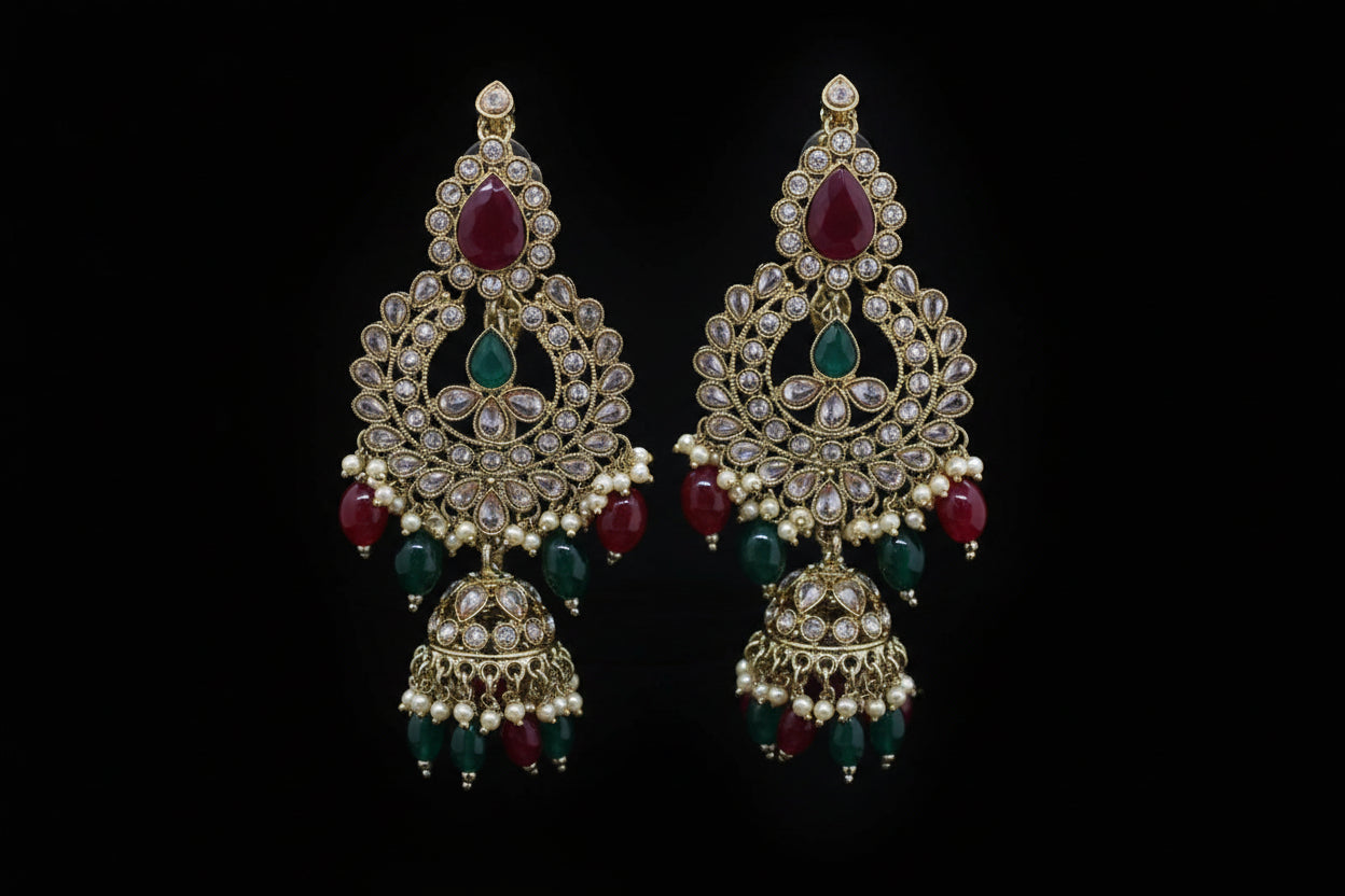 Ornate earrings with red and green stones on a gray stand against a dark background, branded 'SAHELI'.
