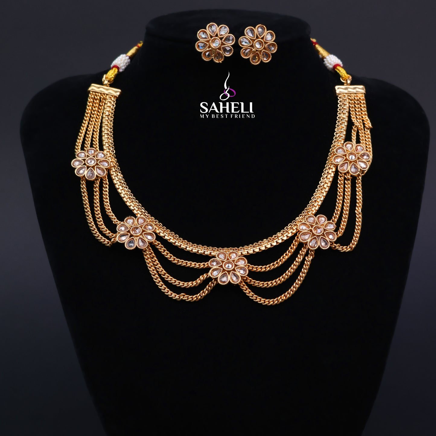 Gold necklace and earrings set on a black stand with 'SAHELI' branding.